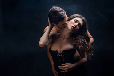 Shirtless man kissing in neck sexy woman in corset on black background with smoke clipart