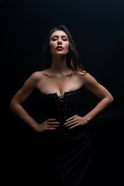 Sexy woman in corset with hand on hips looking at camera isolated on black clipart