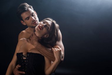 Boynundan tutulan kaslı adam. Korseli seksi kadın. Siyahlar içinde. 