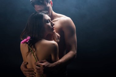Sensual naked woman standing by shirtless boyfriend with gerbera on black background with smoke  clipart