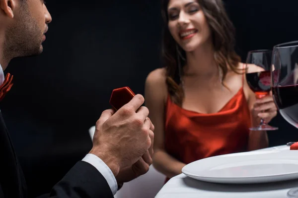 男性の選択的焦点を提示ジュエリーボックスで笑顔のガールフレンドとともにワイングラスロマンチックなディナー中に黒で隔離 — ストック写真