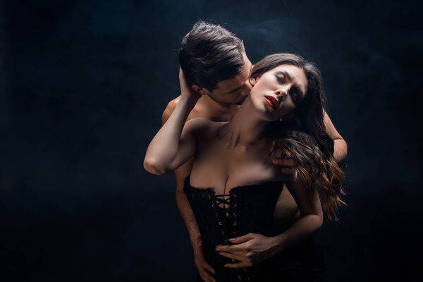 Shirtless man kissing in neck sexy woman in corset on black background with smoke