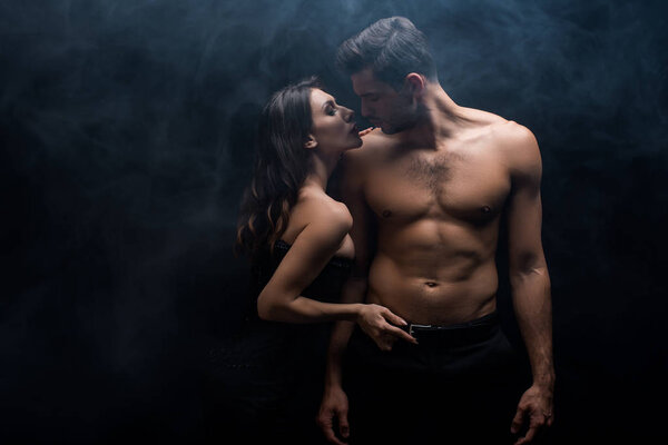 Seductive woman touching belt of muscular man on black background with smoke