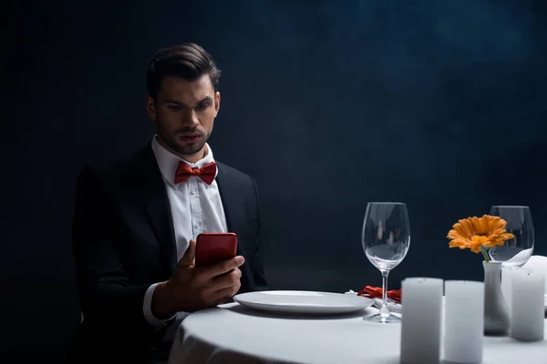 エレガントな男は煙と黒の背景にろうそく付きのテーブルで提供していますスマートフォンを見て — ストック写真