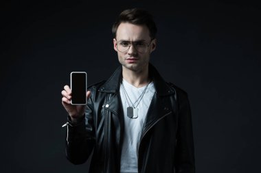 Deri ceketli kaygılı şık adam siyah ekranda boş ekranlı akıllı telefon sunuyor.
