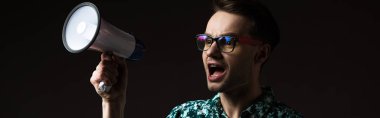 fashionable man in eyeglasses in blue colorful shirt shouting in loudspeaker isolated on black, panoramic shot clipart