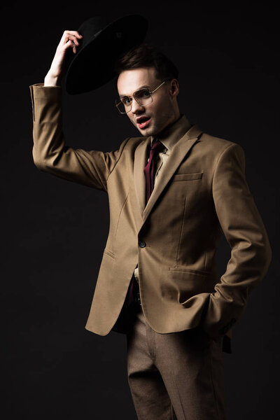 shocked elegant man in beige suit and eyeglasses holding hat isolated on black