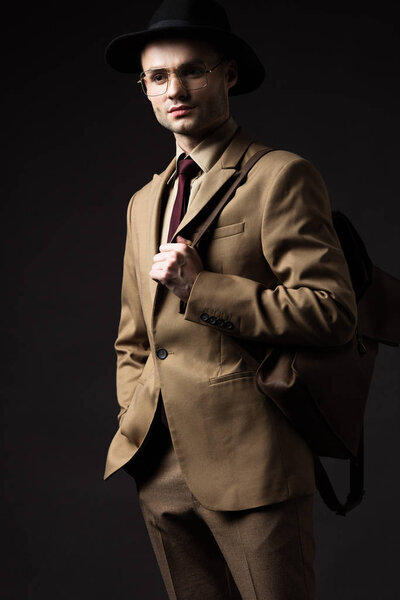 serious elegant man in beige suit, hat and eyeglasses holding brown leather backpack isolated on black