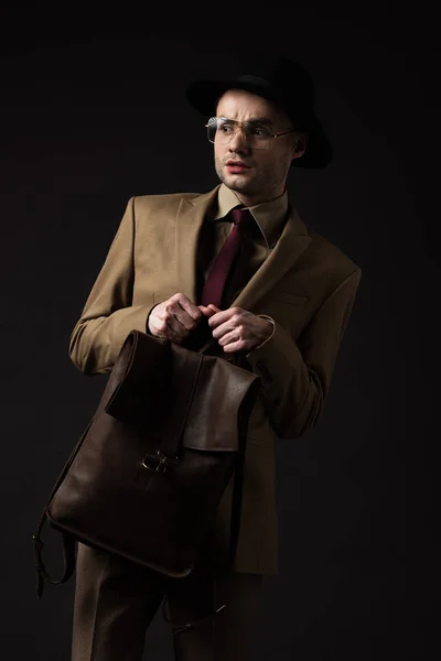 ベージュのスーツに帽子と眼鏡を身に着けているエレガントな男を心配黒で隔離された茶色の革のバックパック — ストック写真