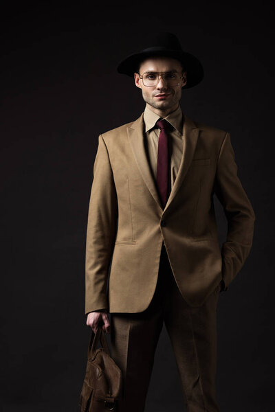 элегантный мужчина в бежевом костюме, шляпе и очках с кожаной сумкой и рукой в кармане изолированы на черном
