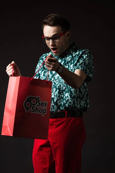 ショックを受けたファッショナブルな男で眼鏡で青カラフルなシャツと赤のパンツ見ますで赤ショッピングバッグからセックスショップ隔離された黒 — ストック写真