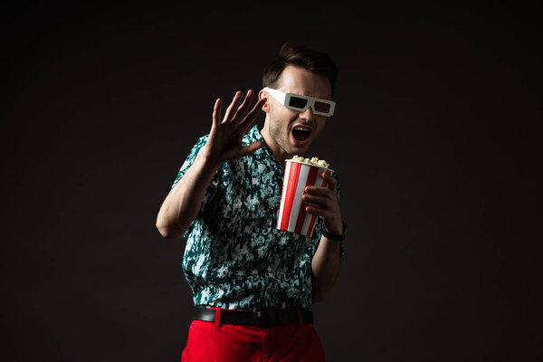 scared fashionable man in 3d glasses in blue colorful shirt and red pants holding popcorn isolated on black
