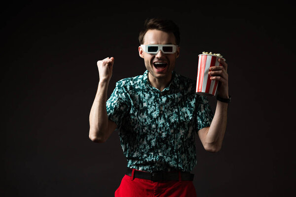 excited fashionable man in 3d glasses in blue colorful shirt and red pants holding popcorn and showing yeah gesture isolated on black