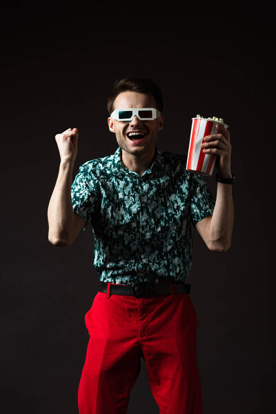 excited fashionable man in 3d glasses in blue colorful shirt and red pants holding popcorn and showing yeah gesture isolated on black