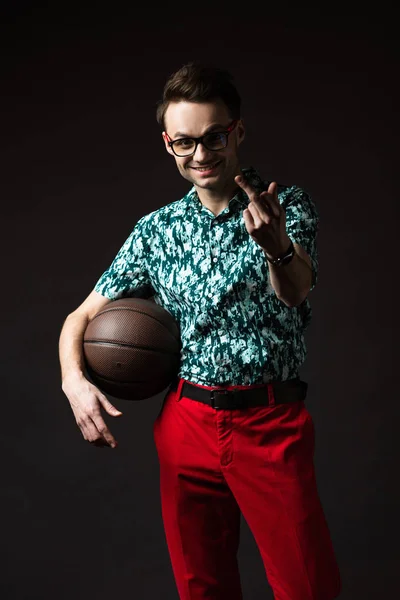 ブルーのカラフルなシャツと赤いパンツを着た眼鏡のファッショナブルな男の笑顔バスケットボールを保持し 黒に隔離された中指を示す — ストック写真