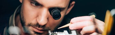 Panoramic shot of watchmaker in eyeglass loupe working with wristwatch isolated on black clipart