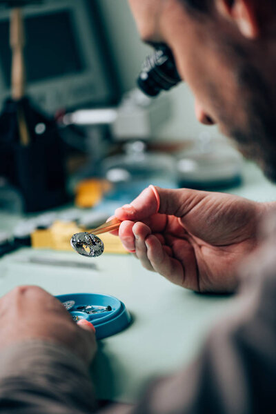 Selective focus of clockmaker in eyeglass loupe holding watch part in tweezers by tool tray on table