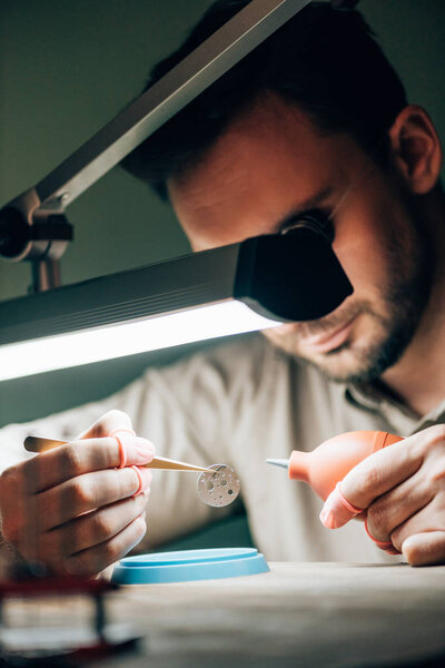Selective focus of clockmaker cleaning watch part with blower by lamp on working table