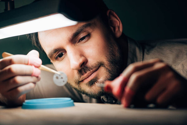 Selective focus of handsome watchmaker looking at clock part by lamp on table