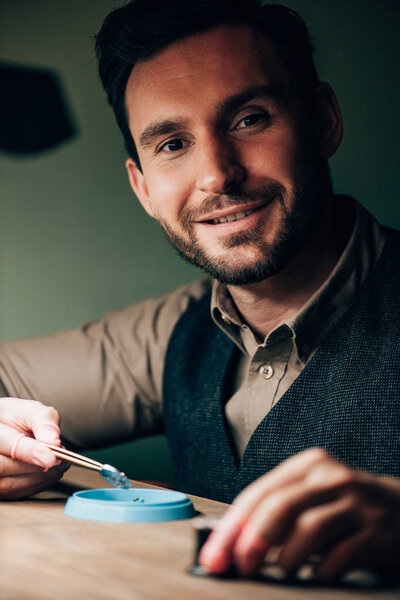 Handsome watchmaker smiling at camera while working with watch parts at table