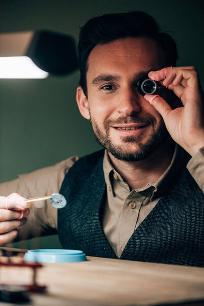 Smiling watchmaker with eyeglass loupe holding watch part while holding at camera