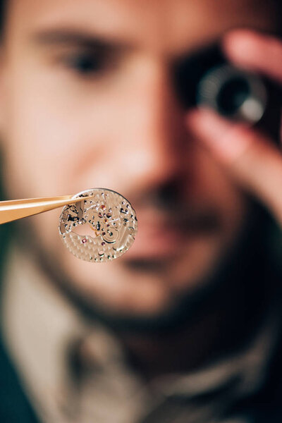 Selective focus of watchmaker holding part of mechanical wristwatch in tweezers 