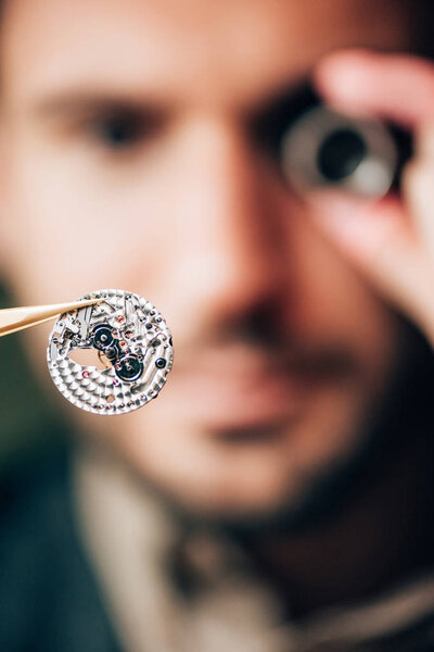 Selective focus of watchmaker holding part of mechanical wristwatch in tweezers
