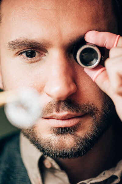 Selective focus of handsome watchmaker holding eyeglass loupe and watch part 