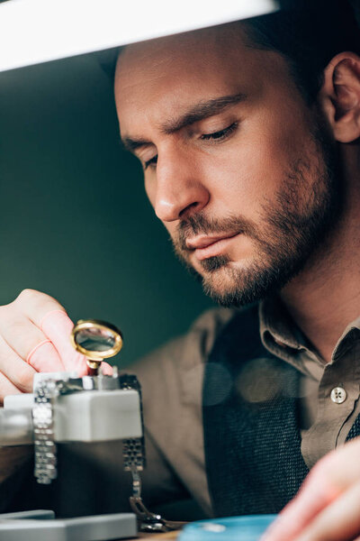 Selective focus of handsome watchmaker working with magnifying glass and wristwatch on movement holder