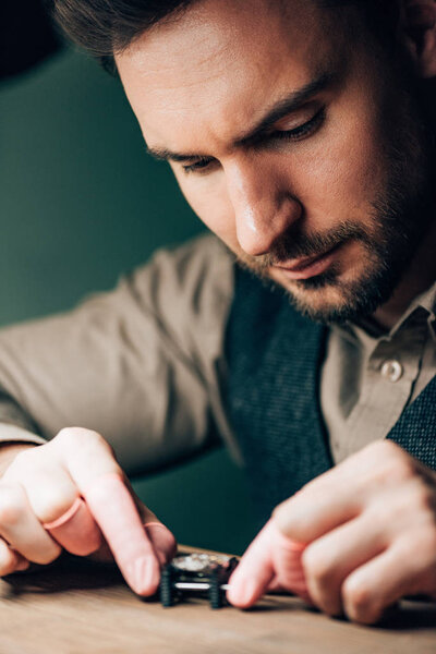 Selective focus of handsome clockmaker working with wristwatch on movement holder on table