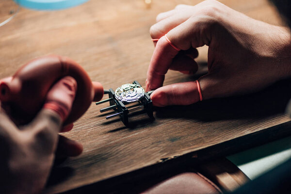 Cropped view of clockmaker using blower for wristwatch on movement holder at table