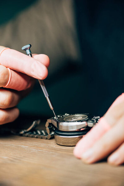 Cropped view of watchmaker in latex finger tips working with wristwatch and screwdriver at table