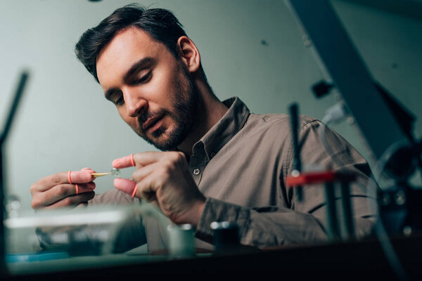 Selective focus of handsome watchmaker working with watch part by equipment on table 