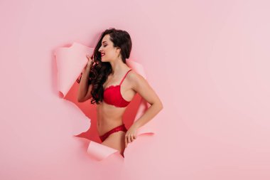 seductive, smiling girl in red lingerie standing in paper hole on pink background clipart