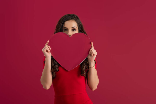 sexy, elegant girl covering face with paper heart while looking at camera isolated on red