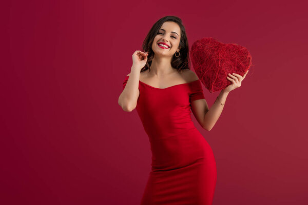 sexy, elegant girl holding decorative heart while smiling at camera isolated on red