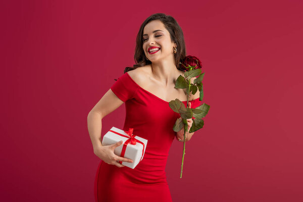 happy, elegant girl holding rose and gift box while smiling with closed eyes isolated on red
