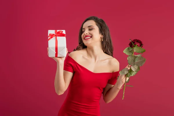 sensual, elegant girl holding rose and gift box while smiling with closed eyes isolated on red