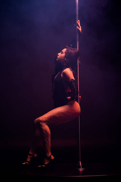 вид сбоку привлекательной стриптизерши, танцующей возле пилона на голубом фоне дыма
 