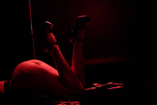 Cropped view of sexy stripper lying near money on black with red lighting