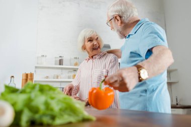 Mutfak masasında dolmalık biber tutarken gülümseyen karısına sarılan yaşlı adamın seçici odağı.