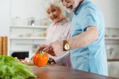 Mutfak masasında karısıyla yemek pişirirken elinde dolma biber tutan gülümseyen adamın seçici odağı.