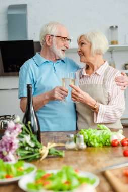 Mutfak masasında şampanya ve sebzelerle kadeh kaldırırken birbirlerine gülümseyen yaşlı çiftlerin seçici odağı.