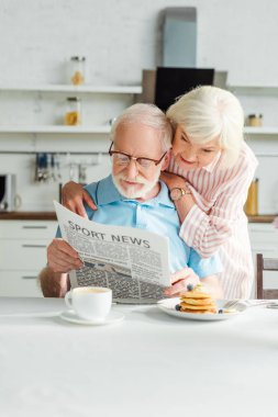 Üst düzey kadın mutfakta kahvaltıda kocasına gazeteyle sarılıyor.