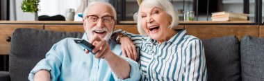 Panoramic shot of senior couple laughing while watching tv on sofa at home clipart