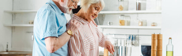 Side view of senior man hugging wife in kitchen, panoramic shot