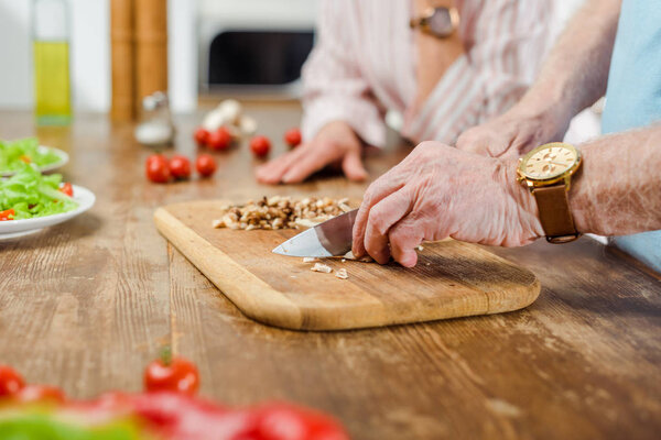 Cropped view of senior man cutting mushrooms beside wife in kitchen