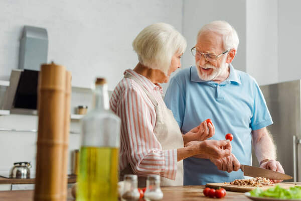 Selective focus of smiling senior man looking at wife while cutting vegetables on kitchen table