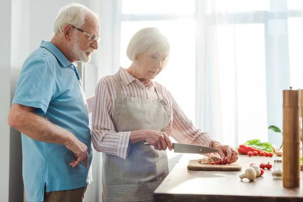 老年人站在妻子身边在厨房桌上切新鲜蔬菜的选择性焦点 — 图库照片