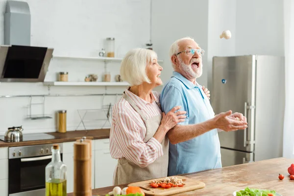 キッチンテーブルの上に新鮮な野菜でキノコを投げる男が笑っているシニアカップル — ストック写真